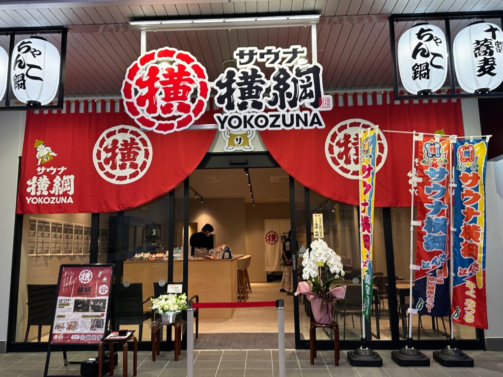 サウナ横綱の入口に掲げられた大きな赤い暖簾と店舗外観。ちゃんこ鍋や十割蕎麦の幟が立つ。