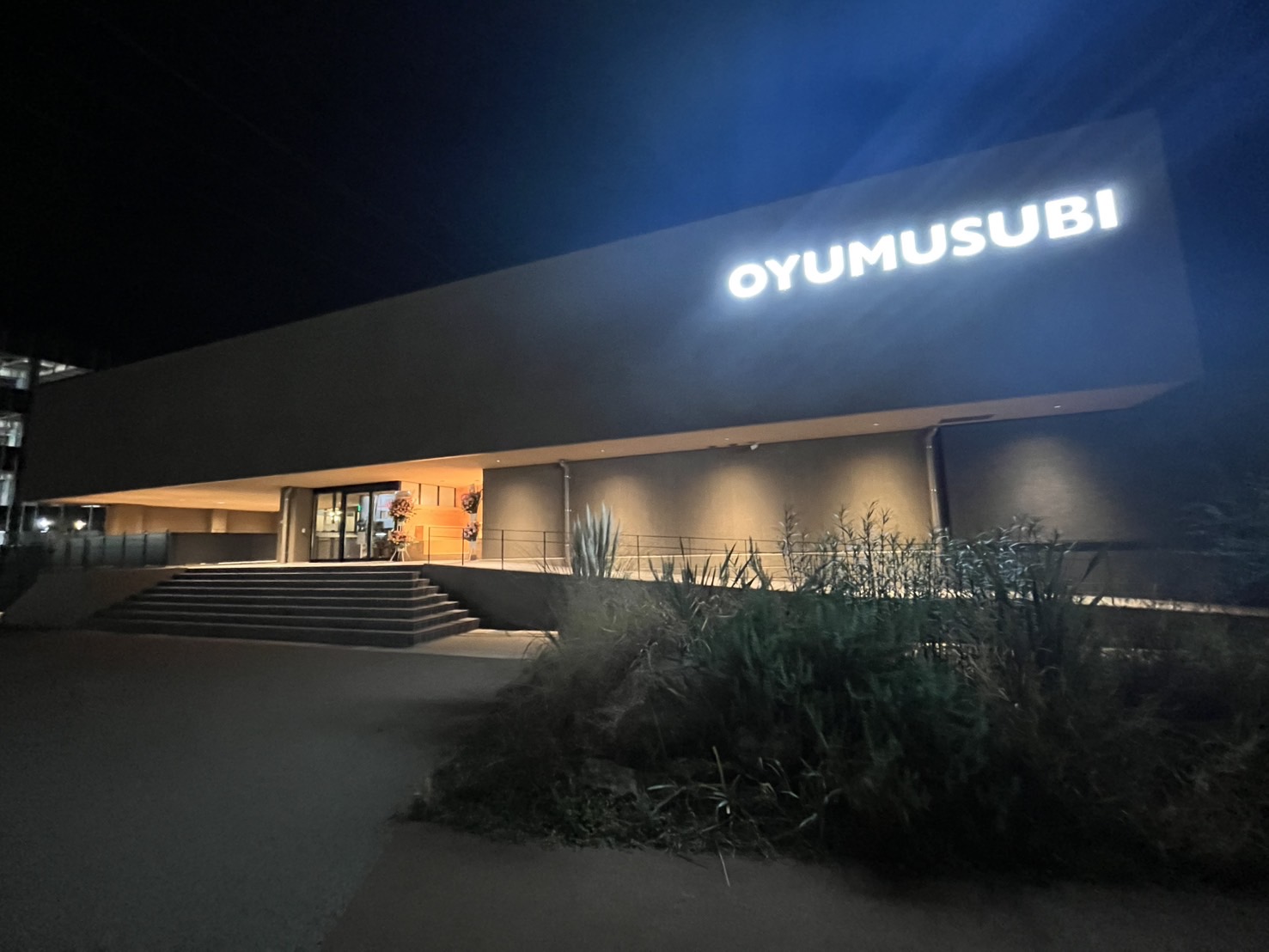 夜間に撮影された「OYUMUSUBI」の正面入口。ライトアップされ、温かい光が周囲を照らしています。