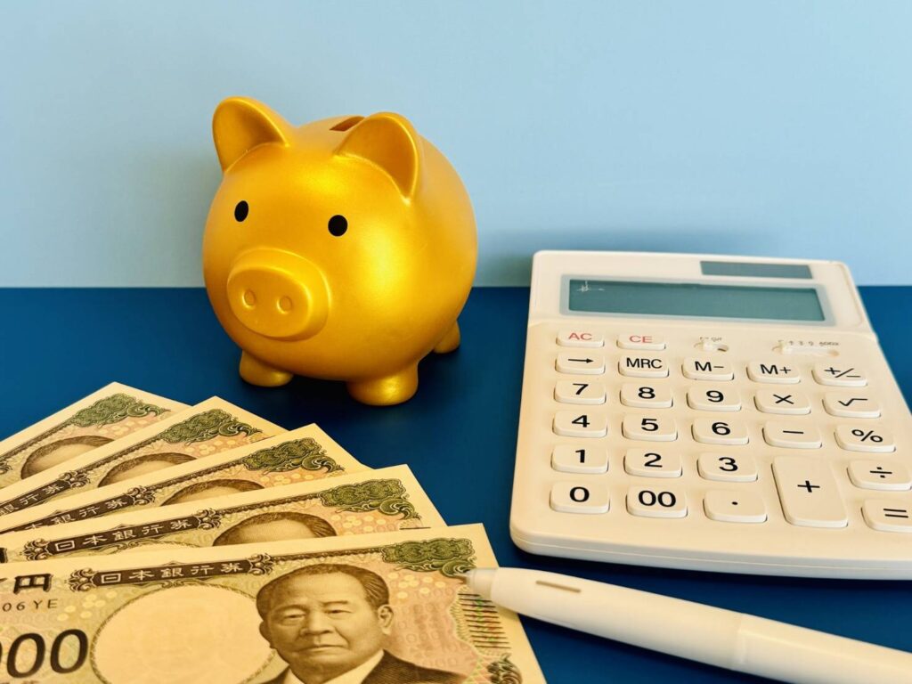 金色の貯金箱と日本円紙幣、電卓、ペンが並んだ節約・資産管理のイメージ