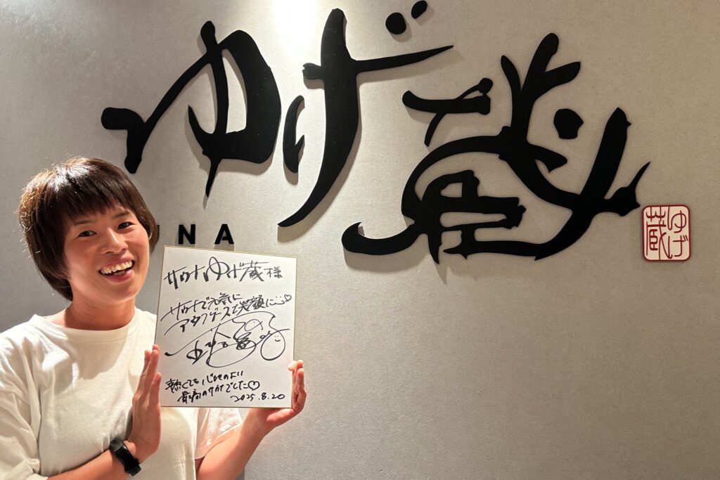 サウナ施設「ゆげ蔵」のロゴ前で色紙を持ち笑顔を見せる五塔熱子さん