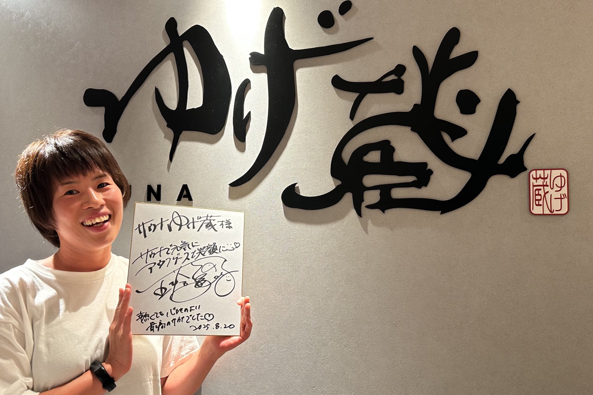 サウナ施設「ゆげ蔵」のロゴ前で色紙を持ち笑顔を見せる五塔熱子さん