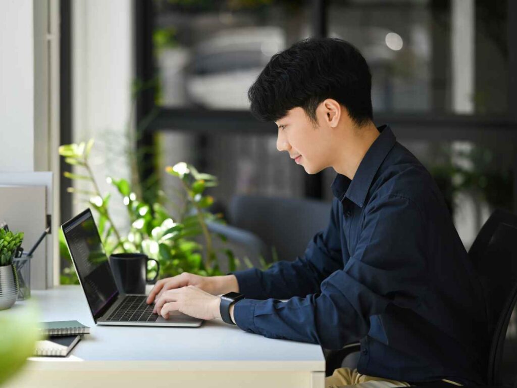 自然光差し込むオフィスでノートパソコンを操作しWeb集客用ホームページを更新する若い男性