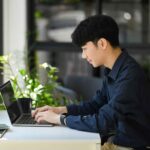 自然光差し込むオフィスでノートパソコンを操作しWeb集客用ホームページを更新する若い男性
