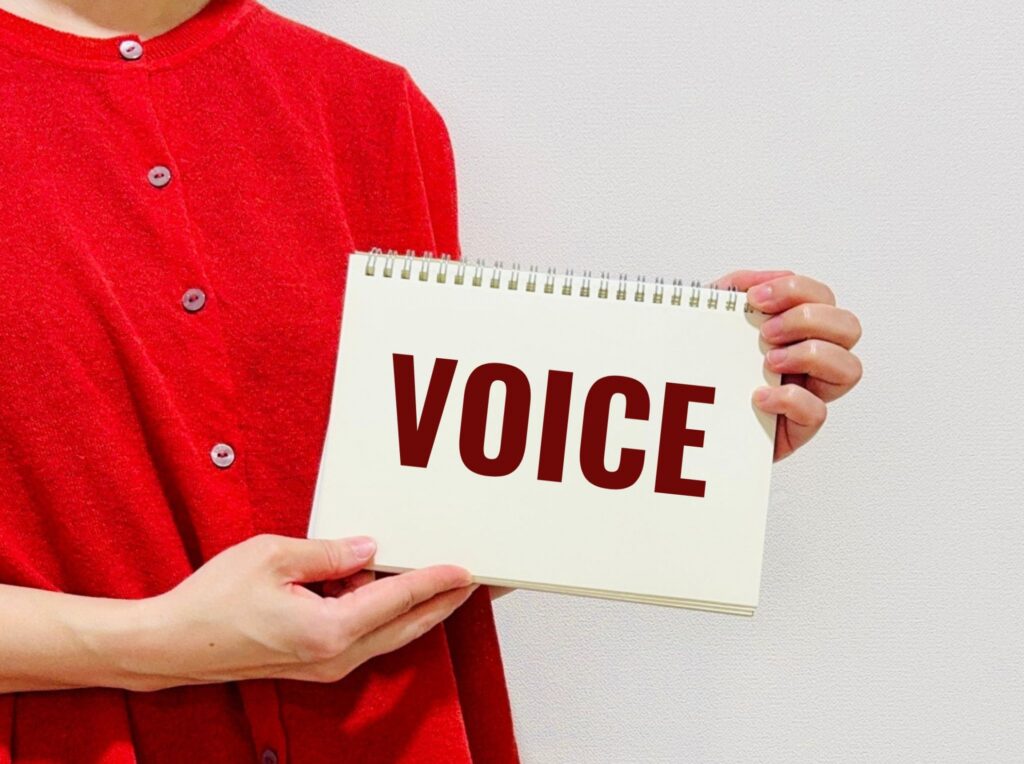 赤い服の人物が「VOICE」と書かれたスケッチブックを正面に掲げている様子