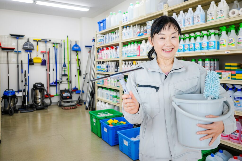 清掃用品が並ぶ店内で笑顔のスタッフがバケツと掃除道具を持つ様子