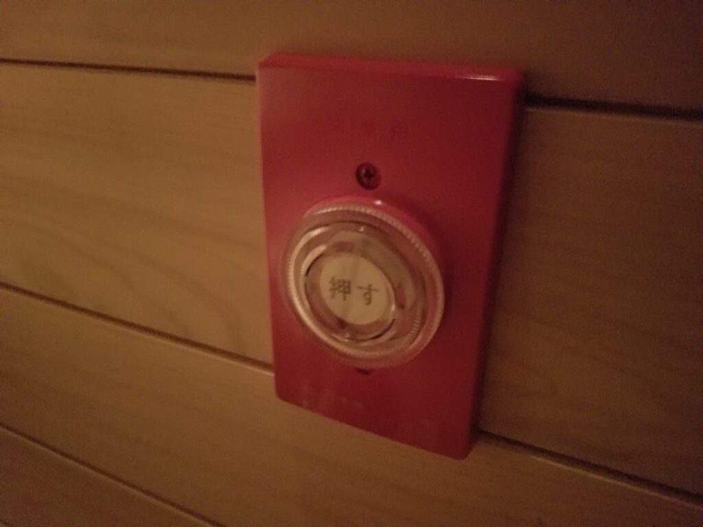 木製パネルの壁面に設置された赤色の非常用押しボタンが写っているサウナ室内の様子。