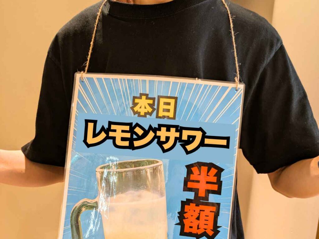 黒いTシャツを着たスタッフが首から「本日 レモンサワー 半額」と書かれたPOPを下げて立っている様子