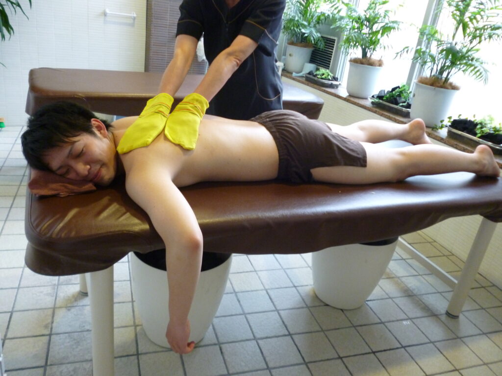 施術台の上でうつ伏せになった男性客の背中を、施術スタッフが手袋を着用してアカスリ施術を行っている様子。