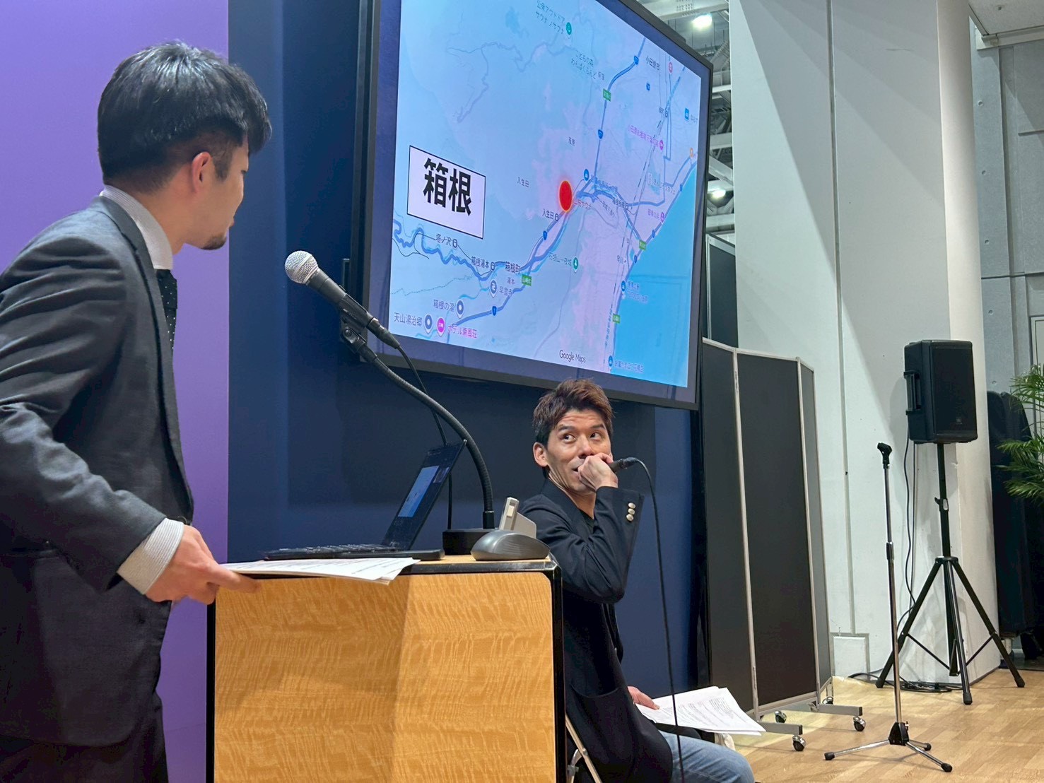 会場の壇上でマイクを持つ男性と、背後のスクリーンに地図が表示されたセミナー風景。
