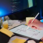 タブレットに表示されたタスク管理の図をスタイラスペンで書き込む様子で、デスク上に付箋とモニターが置かれている。