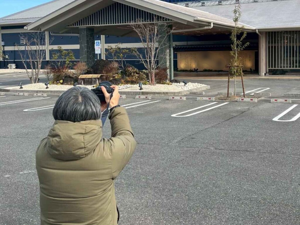 温浴施設の建物外観を駐車場からカメラで撮影している様子で、ホームページ用写真撮影の現場風景が写っている。