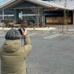 温浴施設の建物外観を駐車場からカメラで撮影している様子で、ホームページ用写真撮影の現場風景が写っている。