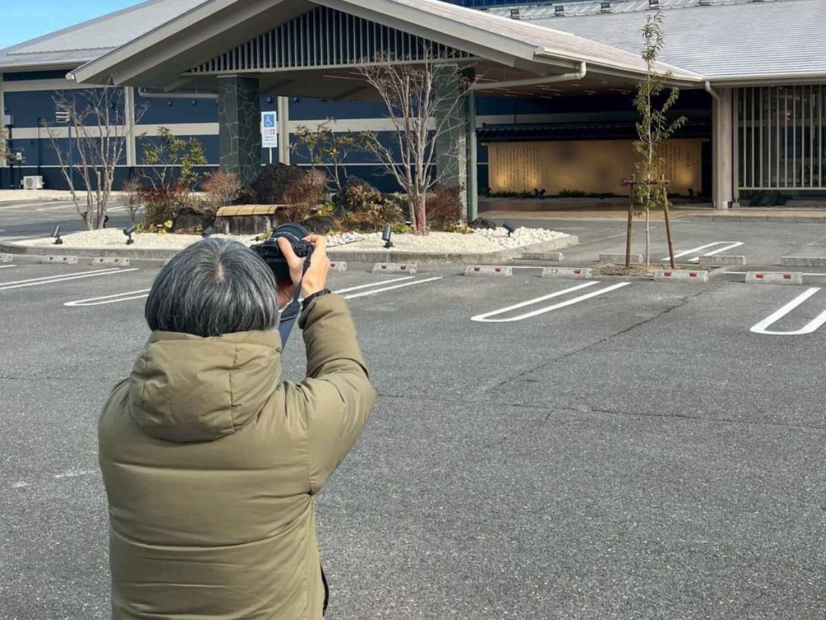 温浴施設の建物外観を駐車場からカメラで撮影している様子で、ホームページ用写真撮影の現場風景が写っている。
