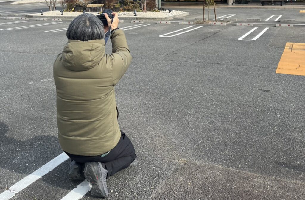 温浴施設の屋外駐車場でカメラを構えて撮影する広報担当者の後ろ姿。