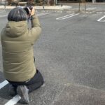 温浴施設の屋外駐車場でカメラを構えて撮影する広報担当者の後ろ姿。