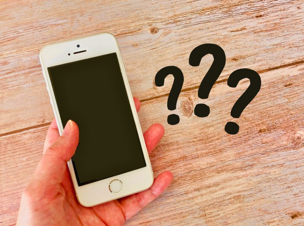 木目のテーブル上で手に持たれたスマートフォンの横に複数のクエスチョンマークが配置されている様子。