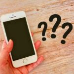 木目のテーブル上で手に持たれたスマートフォンの横に複数のクエスチョンマークが配置されている様子。