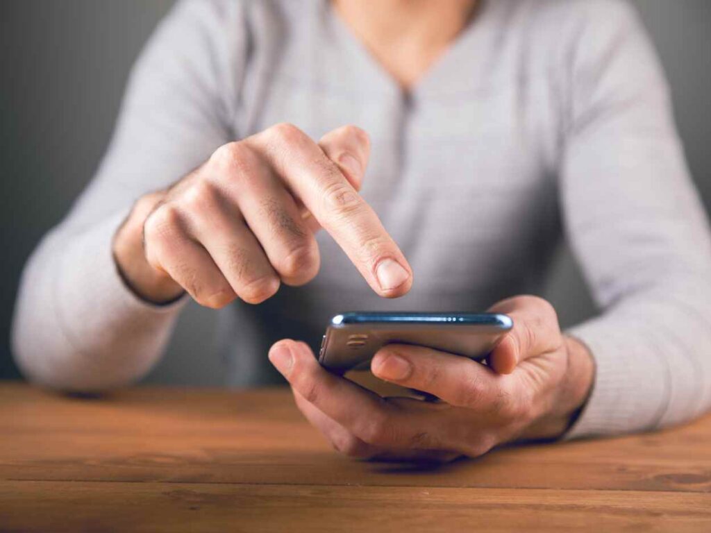 木目調のテーブルの上で、灰色の服を着た人物がスマートフォンを指で操作している様子