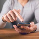 木目調のテーブルの上で、灰色の服を着た人物がスマートフォンを指で操作している様子