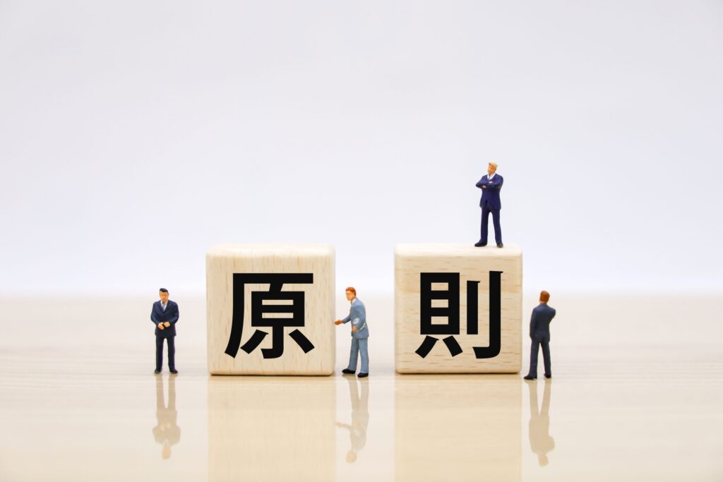 白い背景に「原則」と書かれた2つの木製ブロックが置かれ、その周囲や上にスーツ姿の小さなミニチュア人形が配置されています。