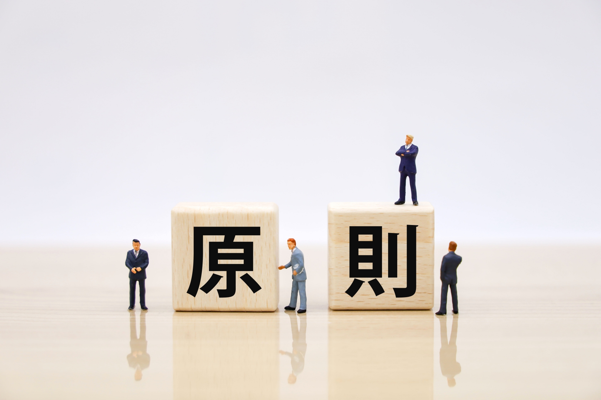 白い背景に「原則」と書かれた2つの木製ブロックが置かれ、その周囲や上にスーツ姿の小さなミニチュア人形が配置されています。