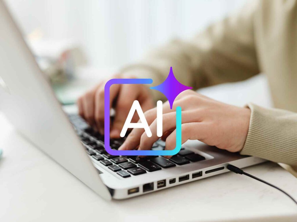 ノートパソコンでタイピングする手元の前面にAIの文字アイコンが重なって表示されている様子。