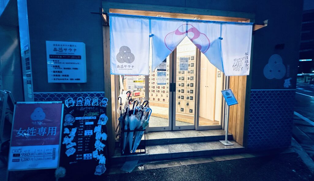 夜の時間帯の品川サウナ入口で、白い暖簾と女性専用日を知らせる看板が設置されています