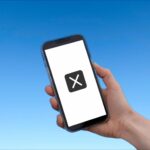 青空を背景にして、画面の中央に大きくバツ印が記されたスマートフォンを手に持っています。