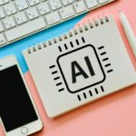 ノートに描かれたAIチップのイラストとキーボード、スマートフォン、ペンが机の上に並んでいるAIイメージ写真です。