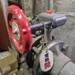 温浴施設の機械室に設置された赤いハンドル付きの配管バルブに、常時開と書かれた管理用の札が取り付けられています。