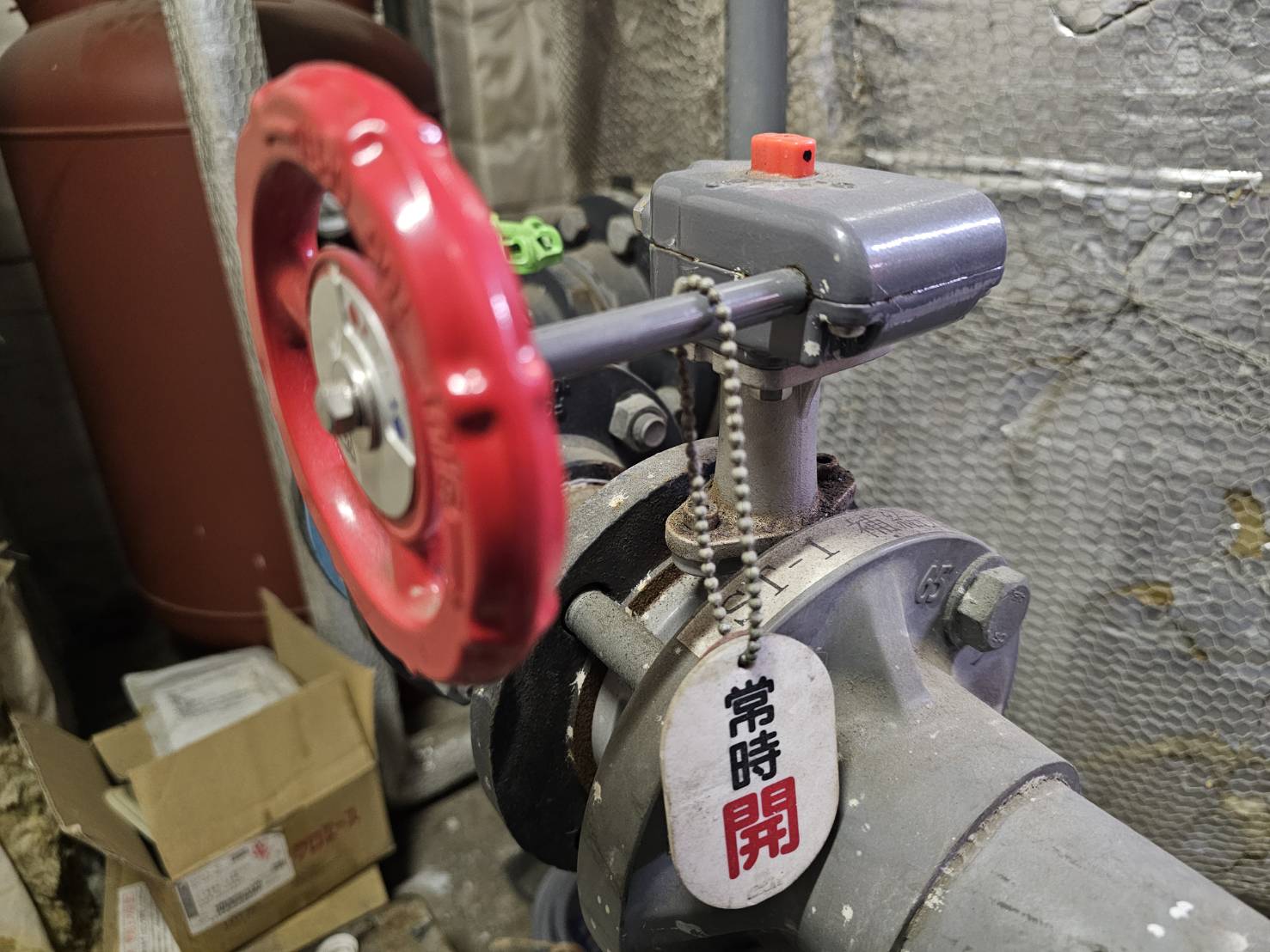 温浴施設の機械室に設置された赤いハンドル付きの配管バルブに、常時開と書かれた管理用の札が取り付けられています。