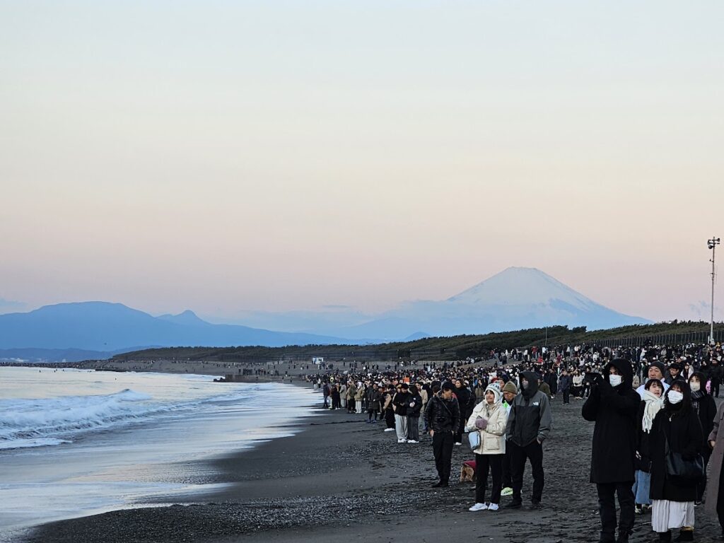 冬の海岸で富士山を背景に日の出を待つ大勢の人々の様子