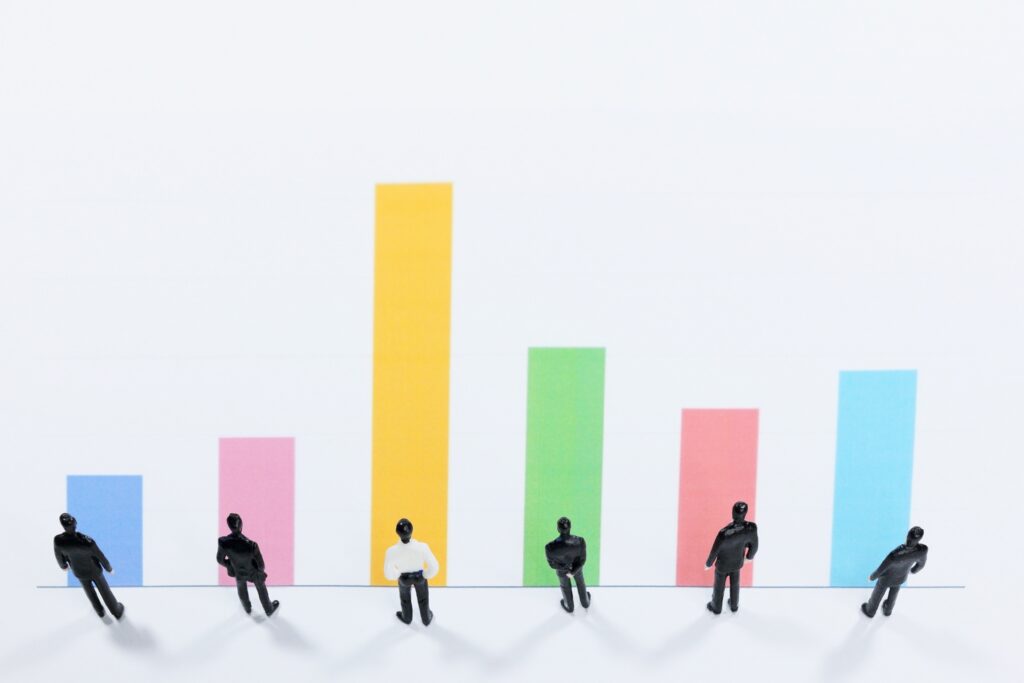 白い背景に整列したビジネスマンのミニチュア人形と、それぞれ異なる高さのカラフルな棒グラフが並んでいます