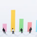 白い背景に整列したビジネスマンのミニチュア人形と、それぞれ異なる高さのカラフルな棒グラフが並んでいます