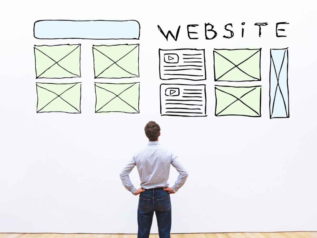 ウェブサイトのワイヤーフレームが描かれた壁の前で、腰に手を当てて構成を検討している男性の背中