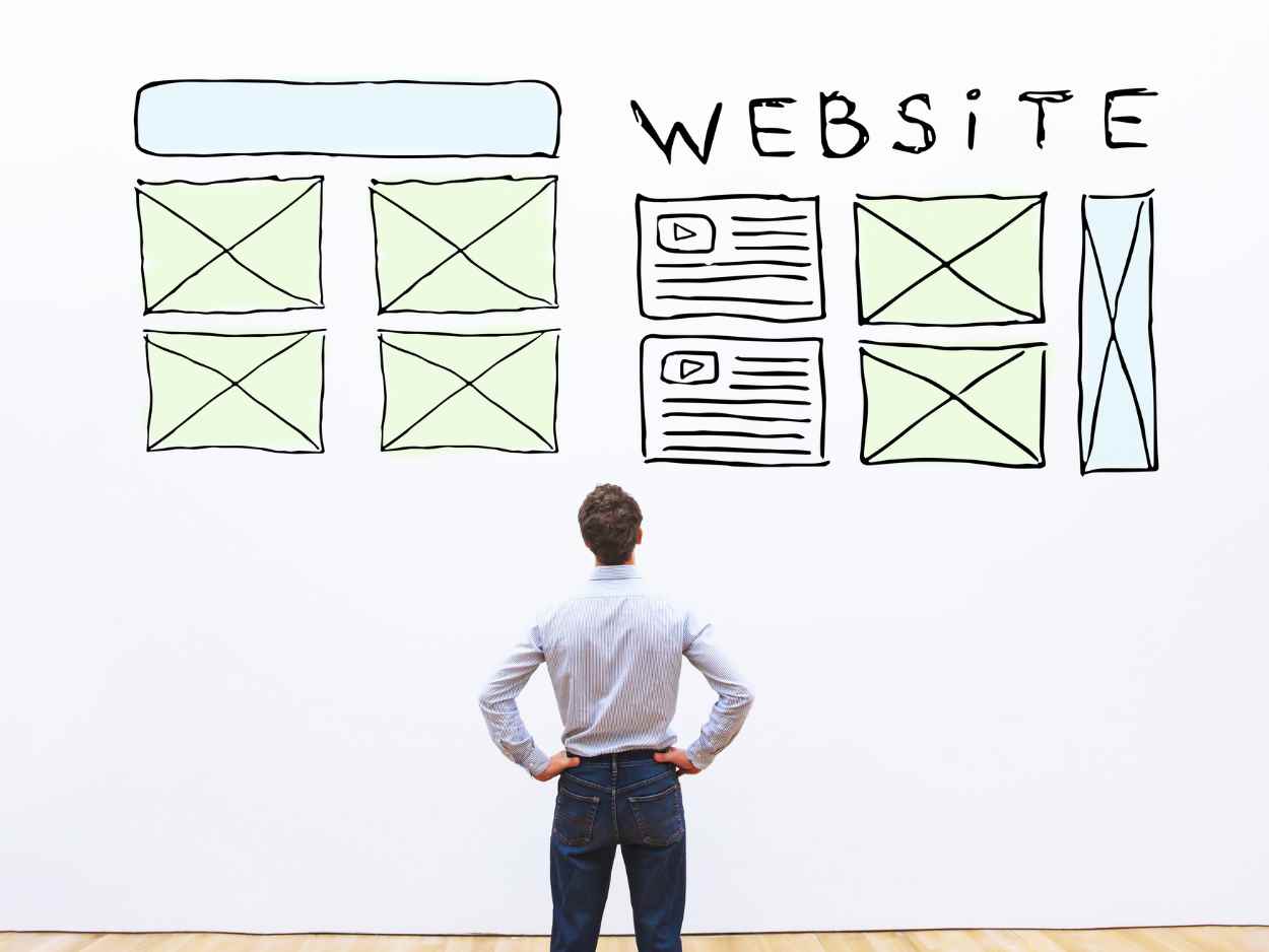 ウェブサイトのワイヤーフレームが描かれた壁の前で、腰に手を当てて構成を検討している男性の背中