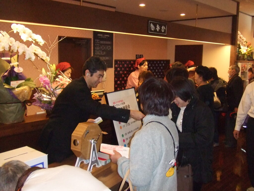 ガラポン抽選会が行われている賑わう温浴施設のフロントで接客するスタッフと客