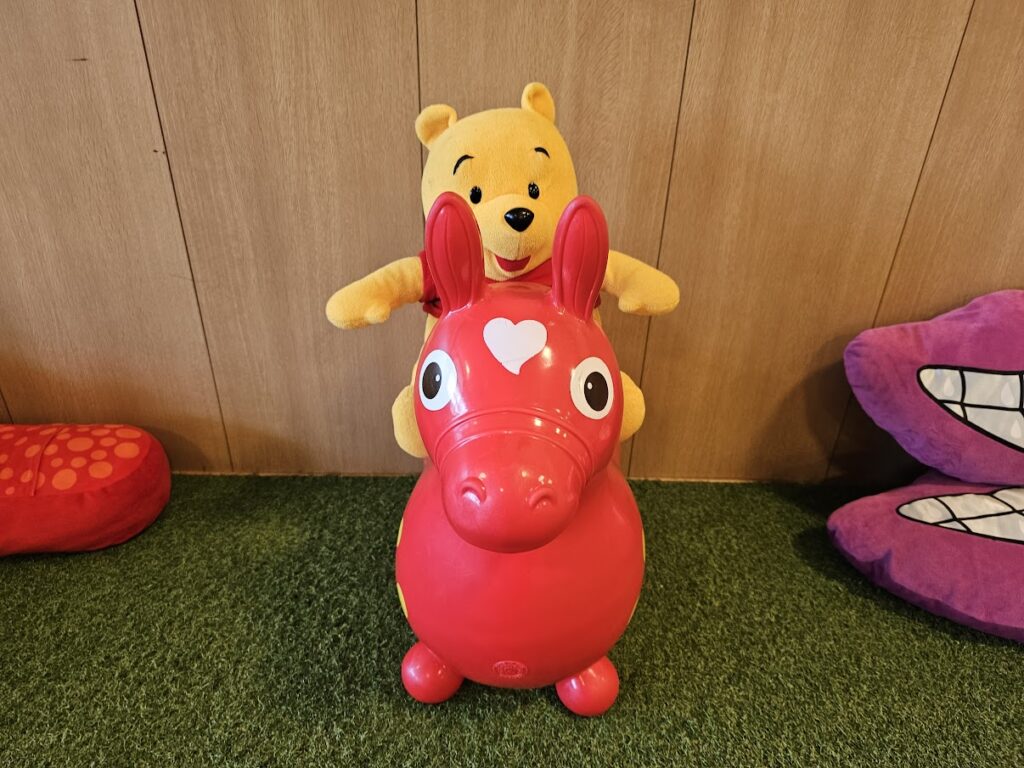 緑色の人工芝の上で、赤い馬型の乗用玩具に乗っているくまのプーさんのぬいぐるみが写っています