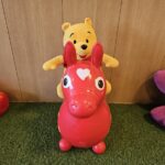 緑色の人工芝の上で、赤い馬型の乗用玩具に乗っているくまのプーさんのぬいぐるみが写っています