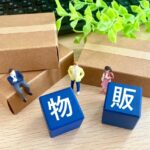 木目調のテーブルの上に置かれた3つの茶色い段ボール箱と、その手前に配置された「物」「販」と書かれた青い立方体、そして3体のミニチュア人形