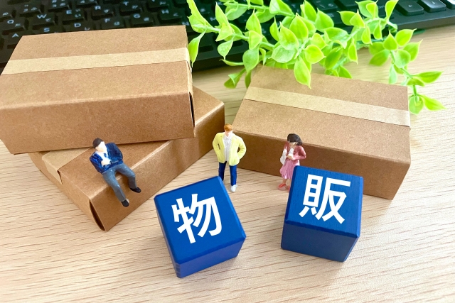 木目調のテーブルの上に置かれた3つの茶色い段ボール箱と、その手前に配置された「物」「販」と書かれた青い立方体、そして3体のミニチュア人形