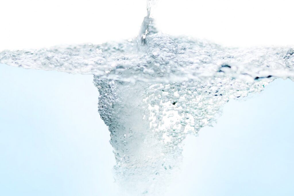 白い背景のなかで、上部から注がれた水が水面に激しい気泡を伴う渦を作り出している様子。