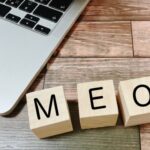 木目調のテーブルの上に置かれたノートパソコンと、MEOというアルファベットが書かれた3つの木製ブロック
