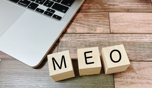 木目調のテーブルの上に置かれたノートパソコンと、MEOというアルファベットが書かれた3つの木製ブロック