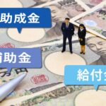 日本円の紙幣の上に、助成金・補助金・給付金と書かれた吹き出しと、スーツ姿の人物のミニチュアが置かれています。