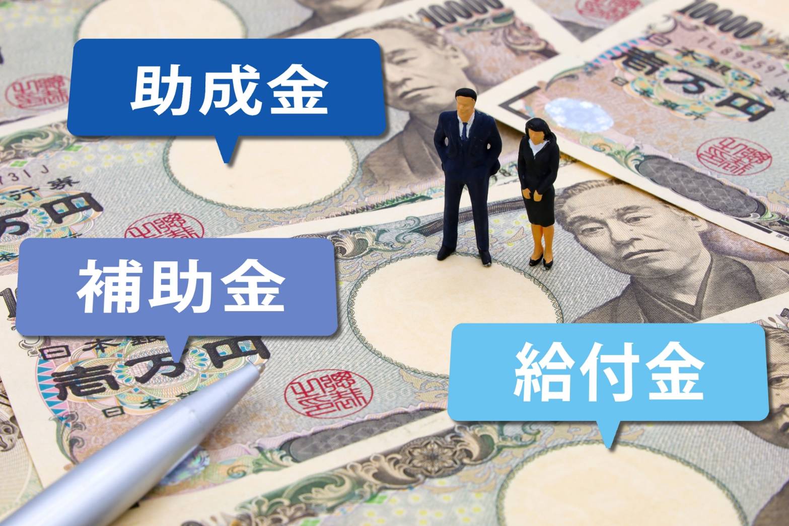 日本円の紙幣の上に、助成金・補助金・給付金と書かれた吹き出しと、スーツ姿の人物のミニチュアが置かれています。