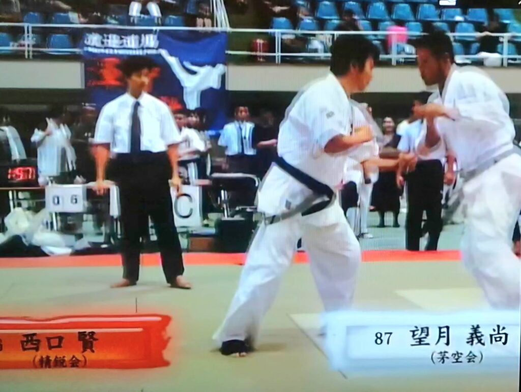 空手の試合で白の道着を着た二人の選手が間合いを取り合い、主審が見守る中で攻防を行う様子