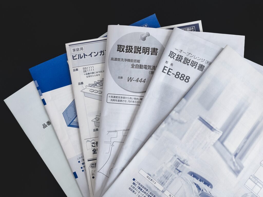 黒い背景の上に、ビルトインガスコンロやオーブンレンジなどの家庭用機器の取扱説明書が複数重ねて置かれています。