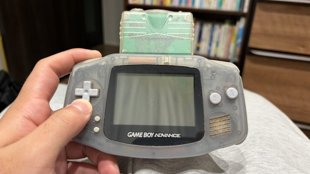 本棚を背景に、緑色のカセットが挿入された透明なゲームボーイアドバンスを手に持つ様子