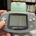 本棚を背景に、緑色のカセットが挿入された透明なゲームボーイアドバンスを手に持つ様子